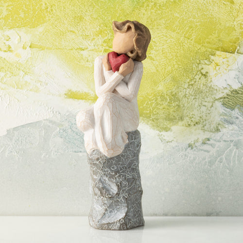 Willow Tree Always, 6”h hand-painted figure in cream dress, knees drawn up to chest, holding red heart to face, seated on gray rock etched with symbols of love, in front of lime painting