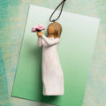 Willow Tree Thank You Ornament, 4”h hand-painted resin figure in cream dress, holding out bouquet of pink peonies, brown elastic cord, laying on green ombre painting