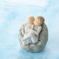Willow Tree My new baby (sky), 2.5”h hand-painted resin figure of toddler in blue onesie holding infant in cream, cuddled together in a beanbag, in front of blue painting