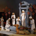 A carved nativity scene set out on a wood table laid with a linen cloth and lit candles in the background.