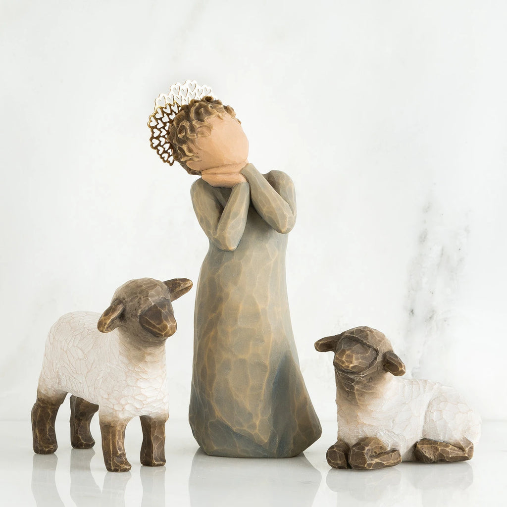 Angel faceless figurine wearing grey dress with hands on her face in between two white lamb figurines
