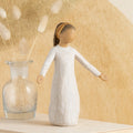 Willow Tree Blessings, 5.5”h hand-painted resin figure in cream dress with darker skin and hair, wearing glitter headband, arms stretched out to sides, on table with pussywillows