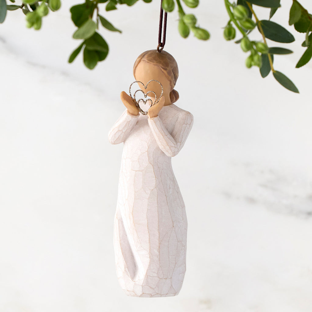 Figure in cream dress, holding thin wire concentric heart in hands. Wire heart is dusted with gold glitter. Hook and loop affixed to figure's head