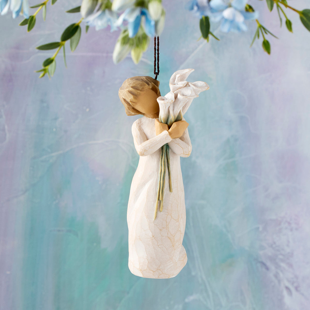 Willow Tree Beautiful Wishes Ornament, 4.5”h hand-painted resin figure in cream dress holding bouquet of white calla lilies, brown elastic cord, hanging in front of blue painting
