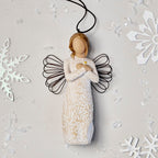 Willow Tree Remembrance Ornament, 4.5”h hand-painted resin angel figure in cream dress carved with symbols of nature, wire wings, crossed hands under small gold-leaf heart, brown elastic cord, laying amongst snowflakes