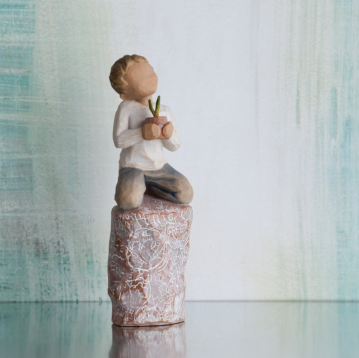 Willow Tree something special, 5.5”h hand-painted figure of small boy holding plant in pot, kneeling on tall rock etched with nature imagery, in front of grey painting