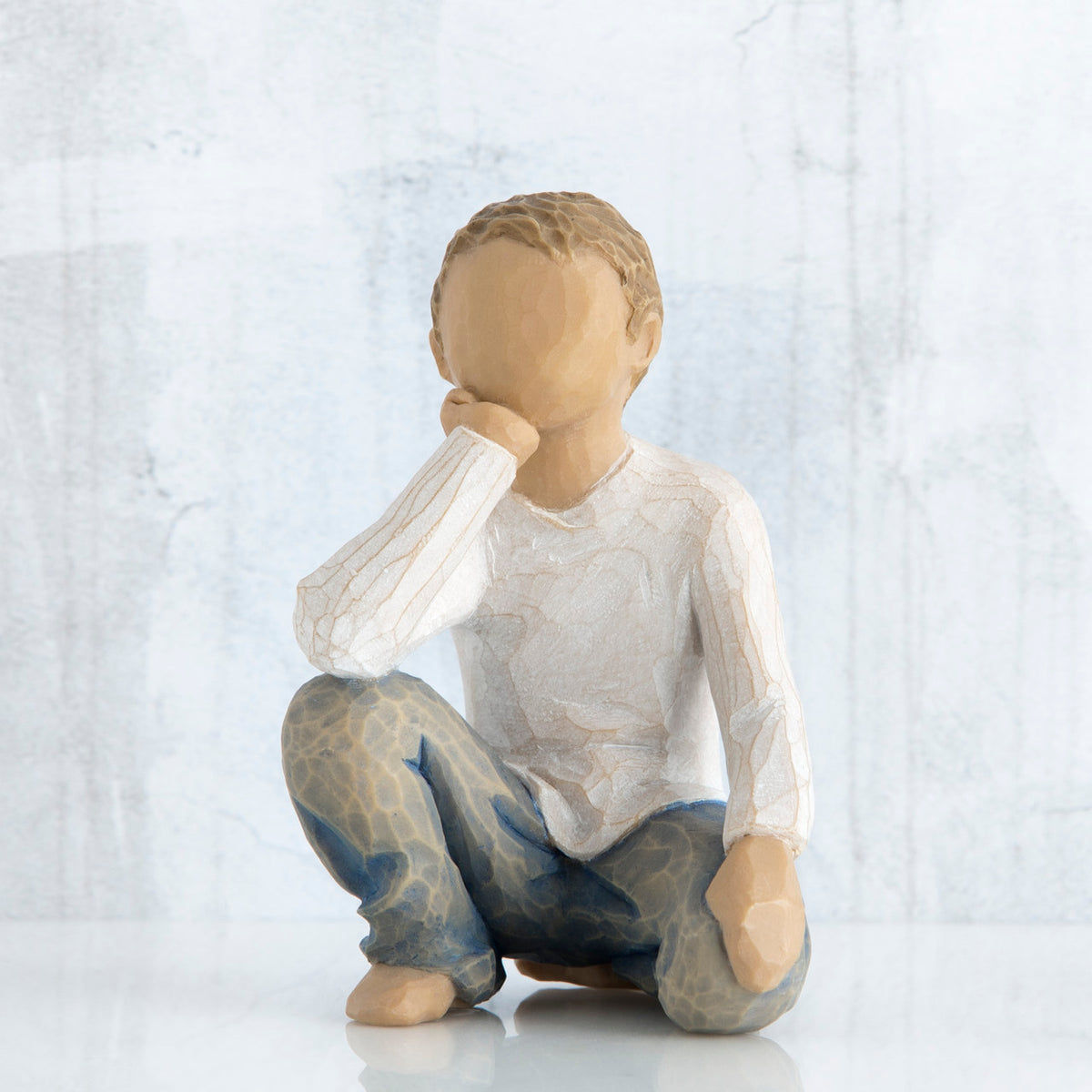 Small faceless boy figurine sitting with elbow on knee - wearing white shirt and blue jeans