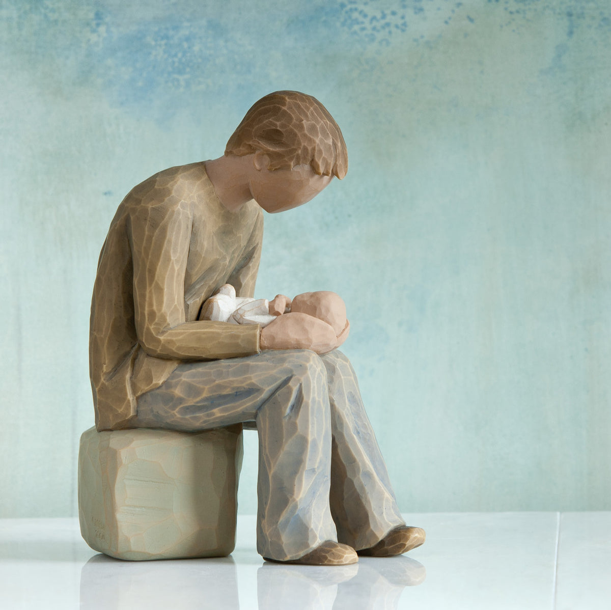 Willow Tree New Dad, 6”h hand-painted seated resin figure in cream shirt and jeans, gazing at newborn infant on lap, in front of teal painting
