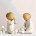 Willow Tree Two Together, two hand-painted seated resin figures of babies, tallest 2", one with hands at mouth, one with hands on legs, both in cream onsies, in front of pastel painting