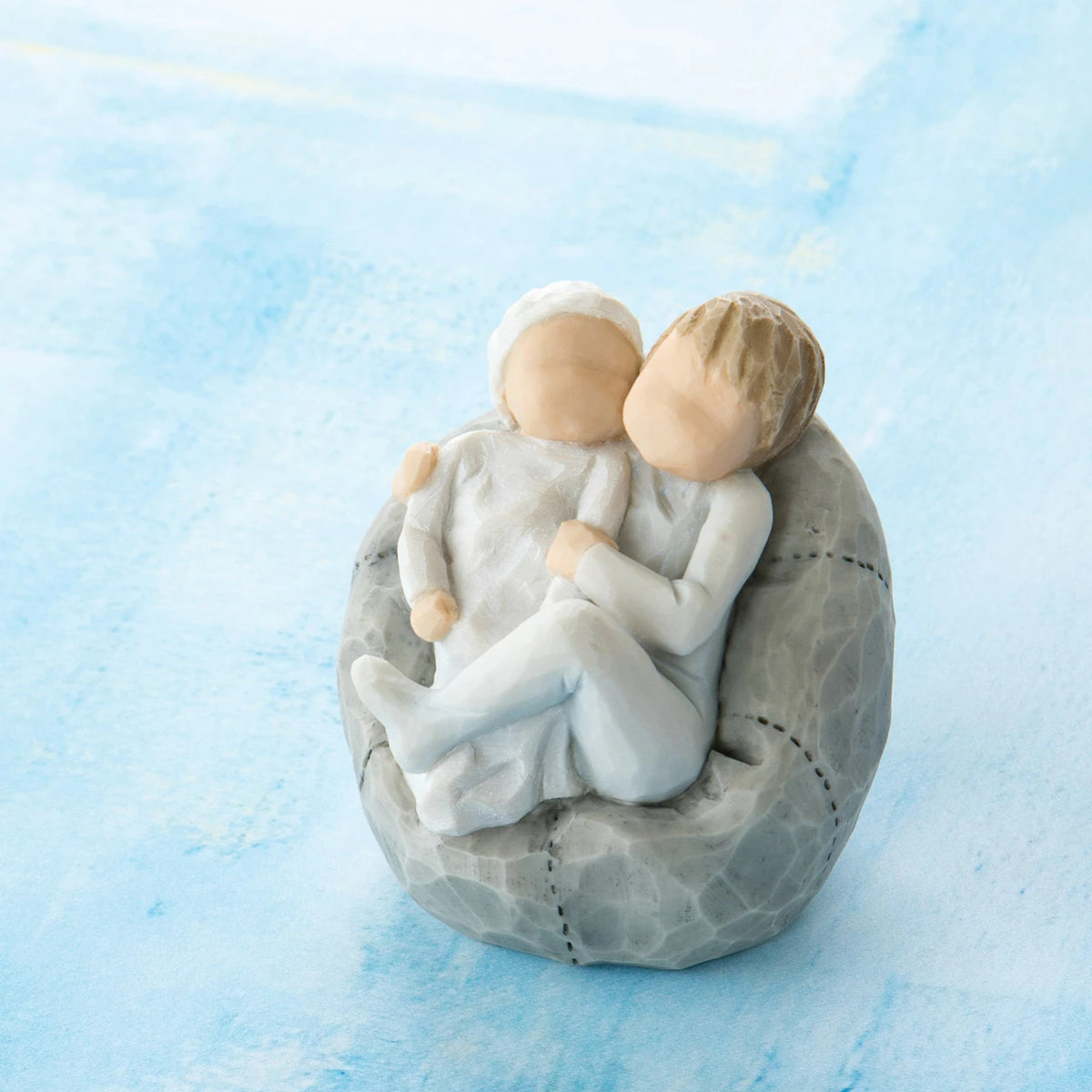Willow Tree My new baby (sky), 2.5”h hand-painted resin figure of toddler in blue onesie holding infant in cream, cuddled together in a beanbag, in front of blue painting