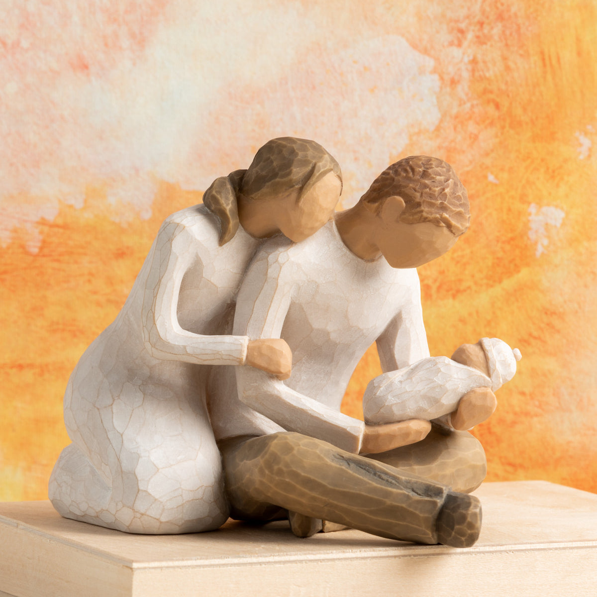 Willow Tree New Life, 5”h hand-painted resin figure of seated couple in neutral colors, holding newborn baby wrapped in cream blanket in front of orange painting