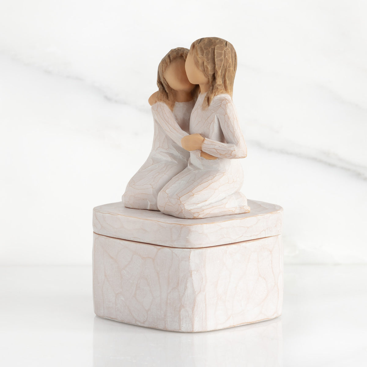 Left side: square resin box, two 3-D figures kneeling atop lid, in cream dresses, hugging each other