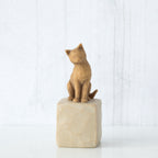 Figure of orange cat with tilted head perched atop a cream colored rectangular pedestal, facing front