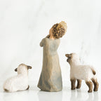 Small girl figurine wearing grey dress facing away from the camera in between two white sheep figurines