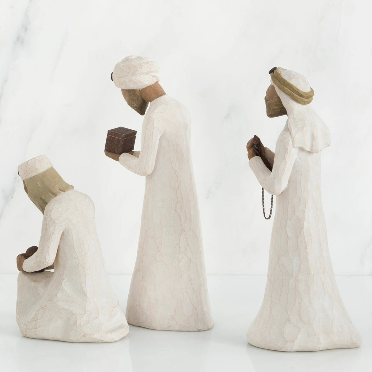 Three wiseman figurines in white dresses facing the side holding brown boxes