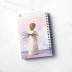 Back view: spiral-bound notebook featuring back of figure with wire wings in cream dress carved with natural symbols, hands crossed under gold heart on chest