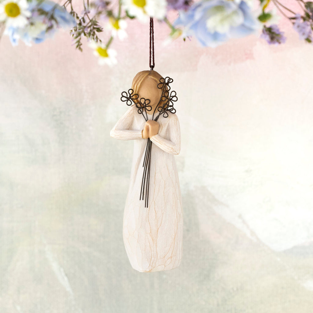 Willow Tree Friendship Ornament, 5.5"h hand-painted resin figure in cream dress holding wire flowers with long stems, brown elastic cord, hanging in front of pastel painting