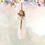 Willow Tree Friendship Ornament, 5.5"h hand-painted resin figure in cream dress holding wire flowers with long stems, brown elastic cord, hanging in front of pastel painting