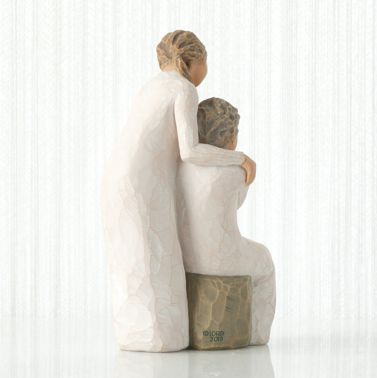 Back view, seated older female in cream dress, with younger female in cream dress, standing at her shoulder, hands embracing