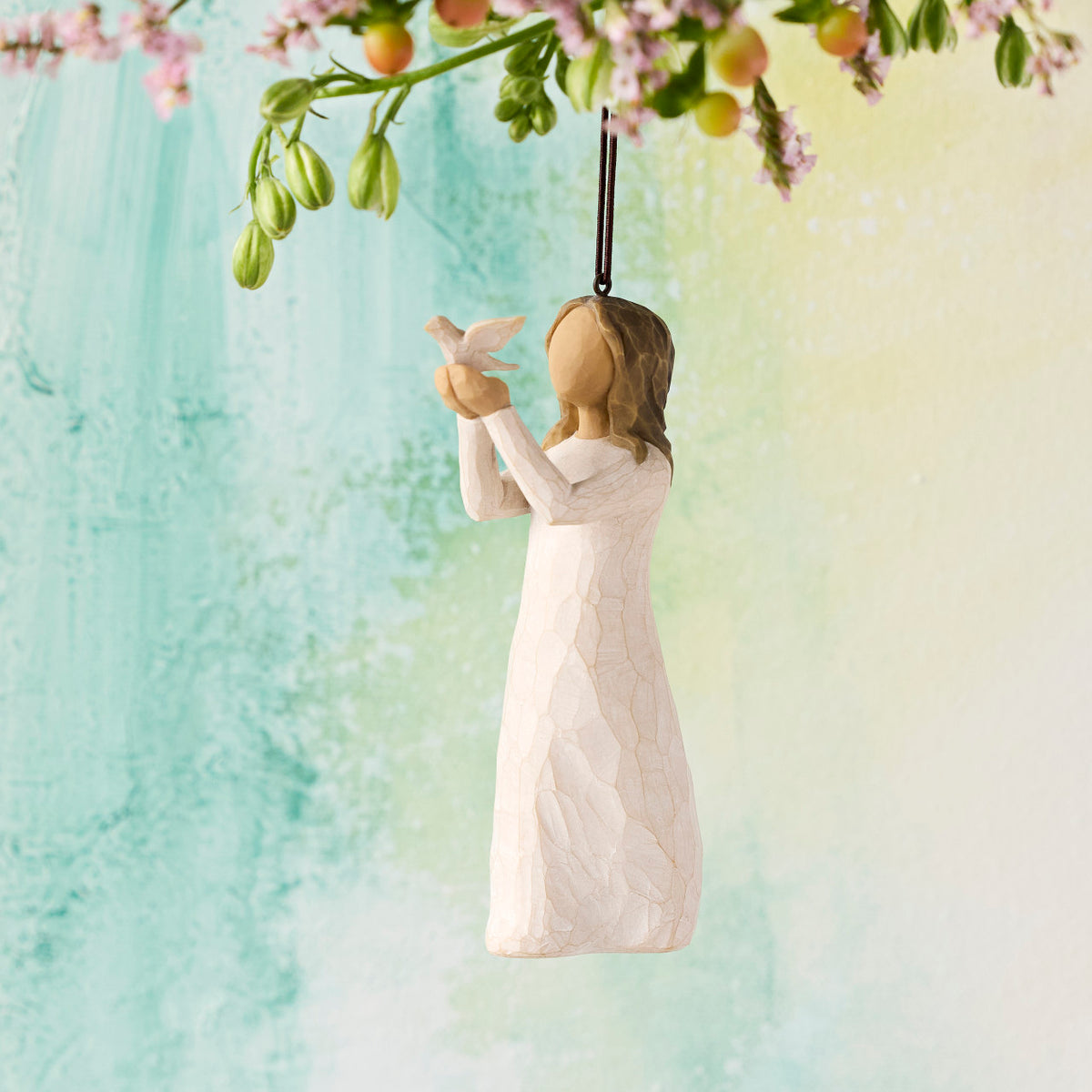 Willow Tree Soar Ornament, 4.5”h hand-painted resin figure in cream dress, holding white dove in extended arms, brown elastic cord, hanging in front of aqua painting