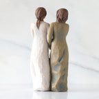 Back view: Figure of two women with darker skin tones in different colored dresses, standing arm in arm