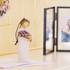 Willow Tree Surprise Ornament 4”h hand-painted resin angel in cream dress, holding large bouquet of purple tulips, brown elastic cord, on table with photos