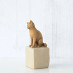 Figure of orange cat with tilted head perched atop a cream colored rectangular pedestal, facing left