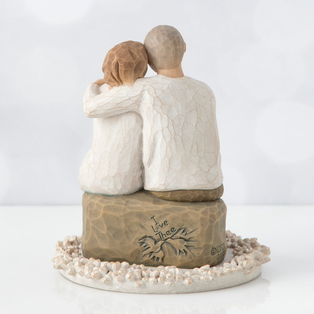 Back view of man and woman figurine in white with arms around another sitting on rock under round white flower plaque