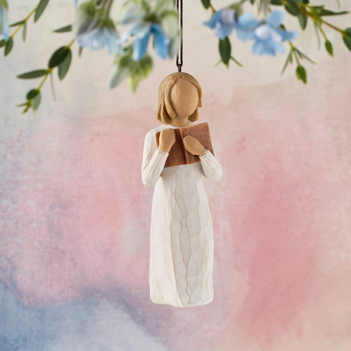 Willow Tree Love of Learning Ornament, 4”h hand-painted resin figure in cream dress holding open book to her chest, brown elastic cord, hanging in front of pink painting