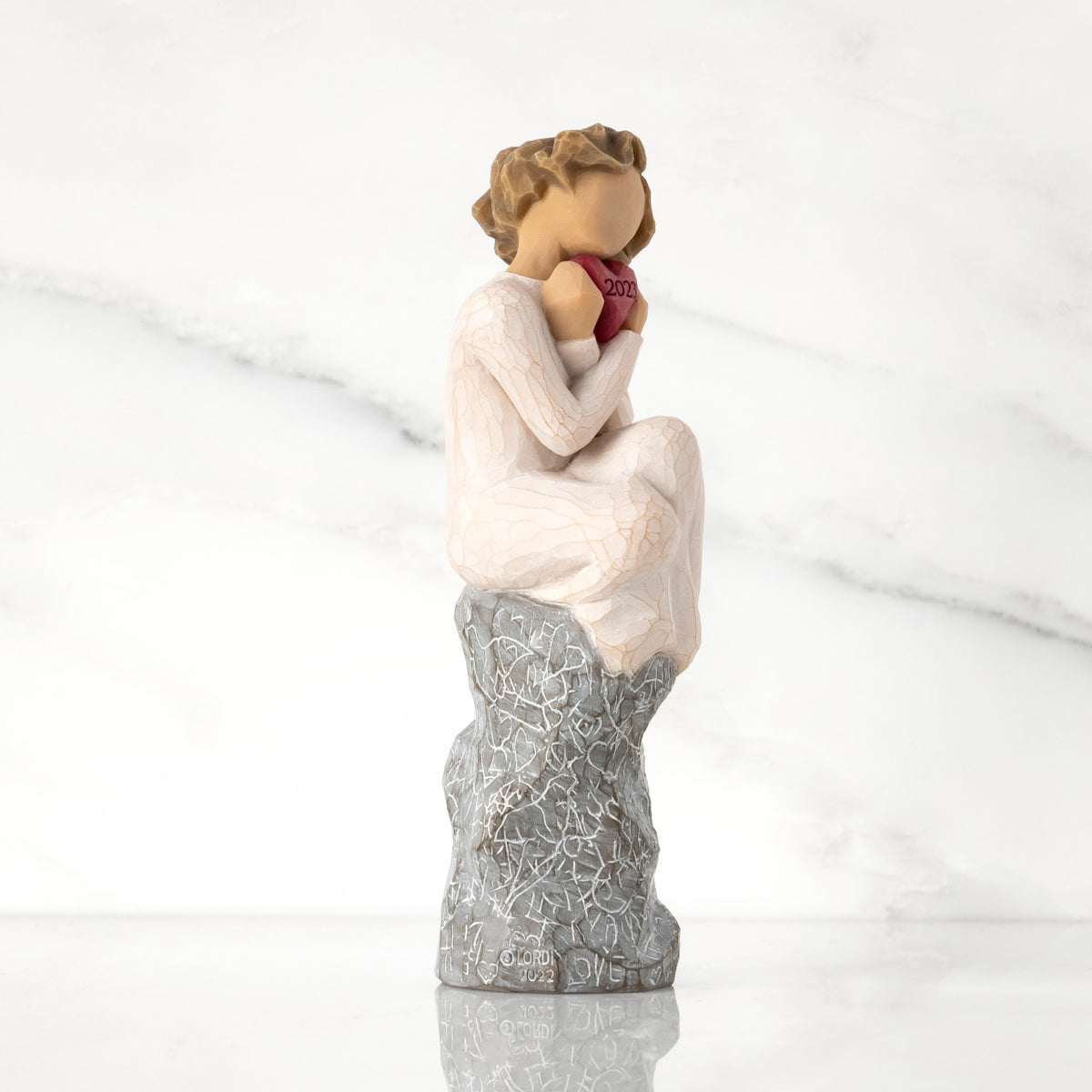 Facing right: figure in cream dress, seated on gray rock, knees drawn up to chest, holding red heart engraved with year 2023