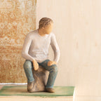 Front view, adult male figure in light gray shirt and blue jeans, seated on gray rock