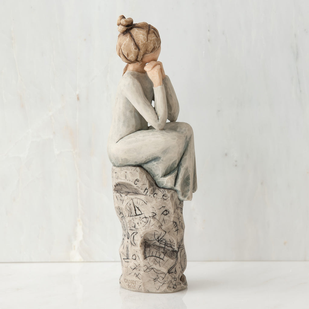 Brunette girl figurine sitting on grey  rock in grey dress with her hands up to her face