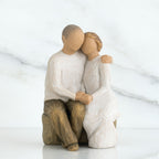 Man and woman faceless figurines hugging one another sitting down on rocl - facing forward - woman is in white, man is in white and brown pants