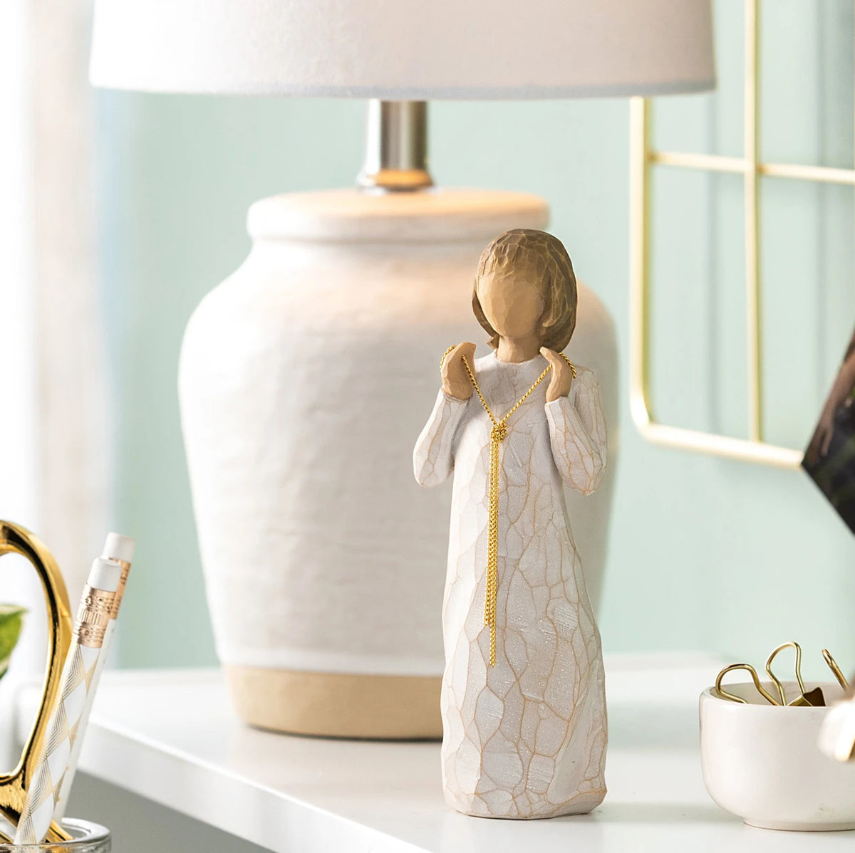 Willow Tree Truly Golden, 5"h hand-painted standing resin figure in cream dress, holding a knotted, long gold chain between her hands, on table with cream lamp