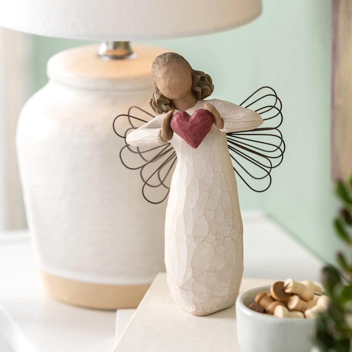 Willow Tree With Love, 5.5”h hand-painted standing resin angel figure with darker skin and hair, in cream dress with wire wings, holding a red heart in hands, on shelf with cream lamp