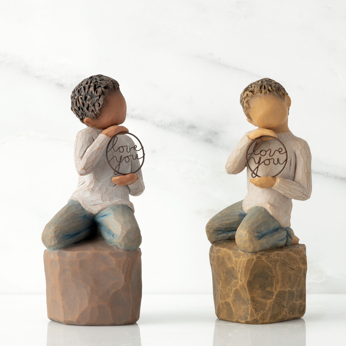 Two options (darker skin and lighter skin) of boy figurine kneeling on rock, holding wire circle with ‘love you’ inside