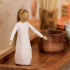 Willow Tree Blessings, 5.5”h hand-painted resin figure in cream dress with darker skin and hair, wearing glitter headband, arms stretched out to sides, on wooden table