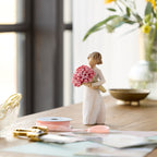 Willow Tree Abundance, 5.5”h hand-painted standing figure in cream dress holding large bouquet of pink roses, on table near window