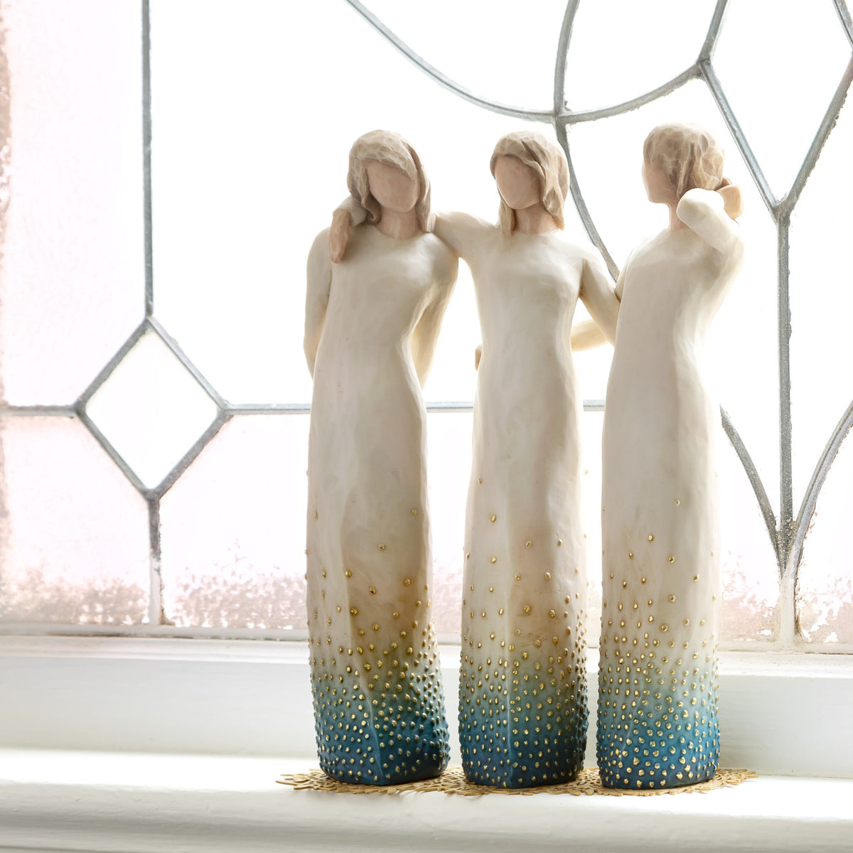 Willow Tree By my side, 9.5" hand-painted resin sculpture of 3 women standing, arms around each other, all in cream and blue ombré dresses with gold-leaf dots, on gold-leaf metal base, by leaded glass window