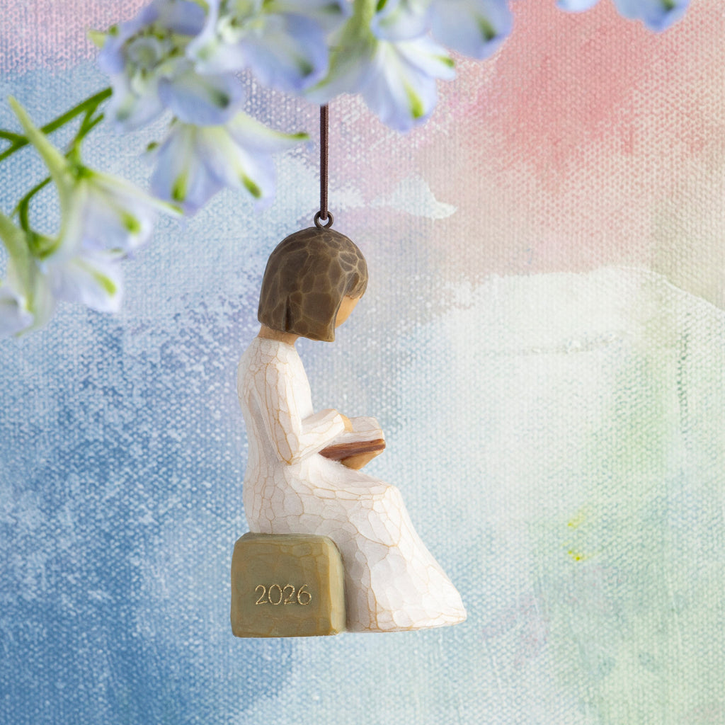 Willow Tree 2026 Ornament, 3”h hand-painted resin figure in cream dress, reading book, seated on rock with 2026 written in gold, brown elastic cord, hanging in front of multi-pastel painting