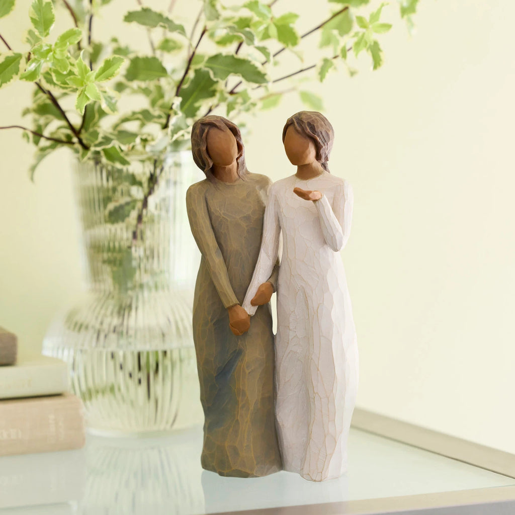 Willow Tree My sister, my friend, 8.5”h hand-painted standing figure of two women with darker skin and hair, standing arm in arm, one in cream dress, one in blue dress, on table with greenery
