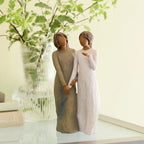 Willow Tree My sister, my friend, 8.5”h hand-painted standing figure of two women with darker skin and hair, standing arm in arm, one in cream dress, one in blue dress, on table with greenery