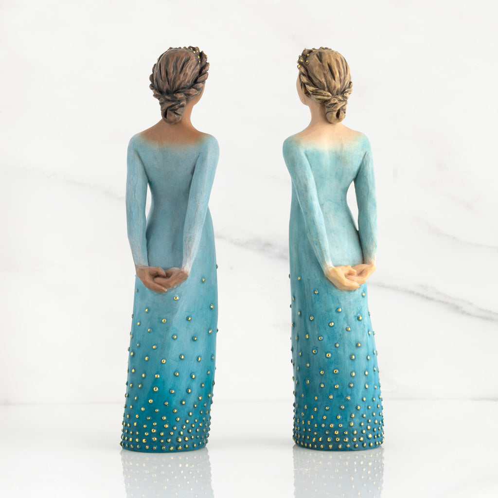 Back view of both lighter and darker-skin figures, arms clasped behind backs, hair and deep blue ombré garments dotted with gold leaf.